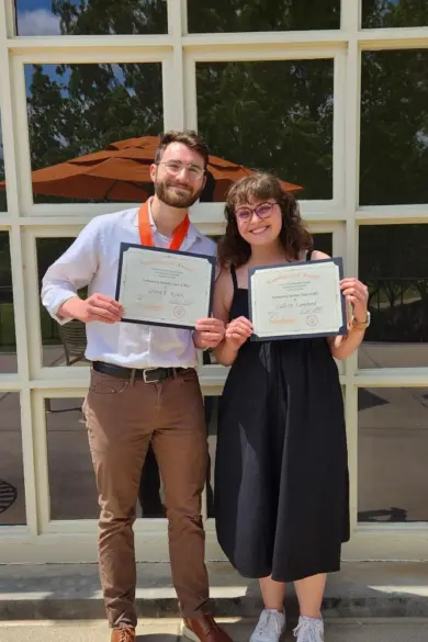 Two people stand smiling in front of a window, each holding a certificate. The person on the left wears a white shirt, brown pants, and an orange ribbon. The person on the right wears glasses and a black dress. There are trees and umbrellas outside the window.