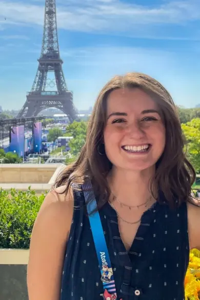 Jillian Michales smiles in front of the Eiffel Tower on a sunny day. They are wearing a sleeveless black top and a blue lanyard, standing amidst green foliage and sunflowers. The sky is clear and blue.