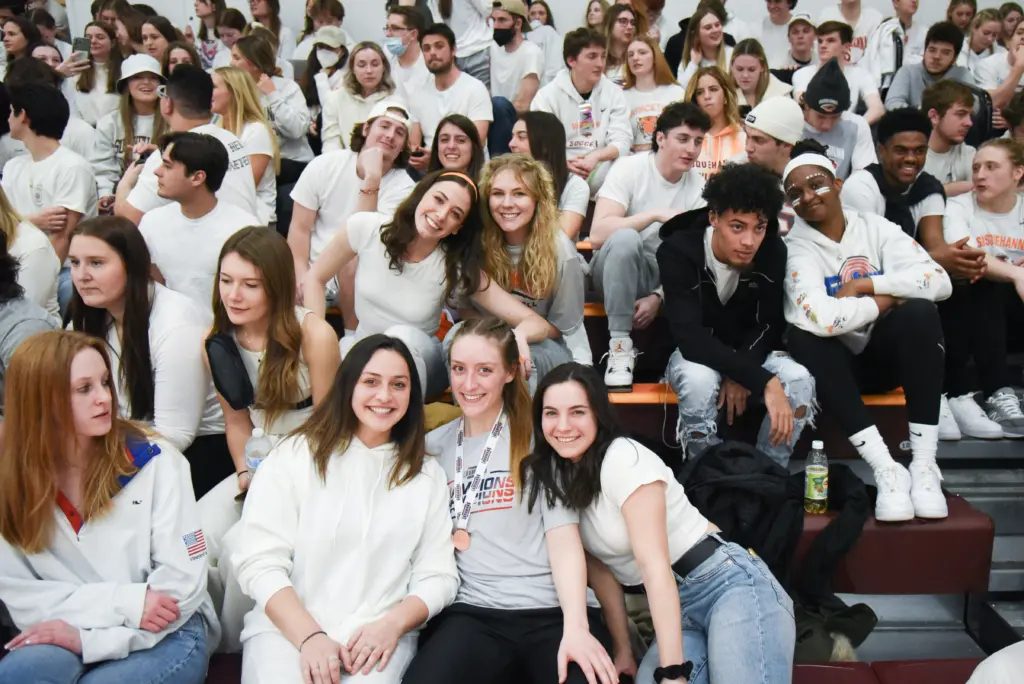 A large group of people are sitting and standing on bleachers, many wearing white shirts. Some are smiling and posing for the camera, while others look around. The crowd appears to be at a spirited indoor event, with a lively atmosphere.