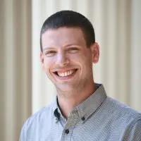 Smiling man with short hair, wearing a light gray button-up shirt, stands against a blurred, light-colored background.