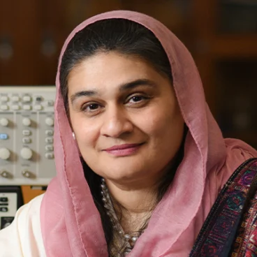 A woman with dark hair, wearing a light pink headscarf and traditional attire, smiles softly at the camera. She is in an indoor setting with electronic equipment in the background.