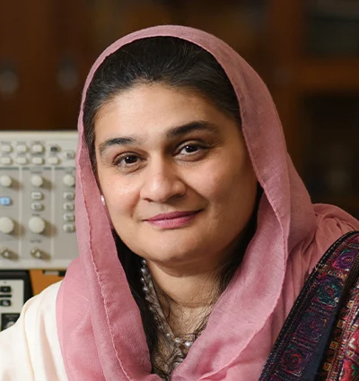 A woman with dark hair, wearing a light pink headscarf and traditional attire, smiles softly at the camera. She is in an indoor setting with electronic equipment in the background.