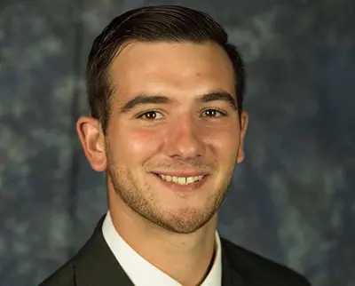 A man in a dark suit and white shirt smiles against a blurred dark blue background. He has short, neatly combed dark hair and light facial hair.