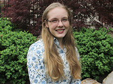A young woman with long blonde hair and glasses smiles while sitting in front of green bushes and a red-leafed tree. She is wearing a floral patterned shirt.