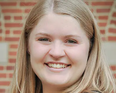 A person with long, light brown hair smiles against a brick wall background.