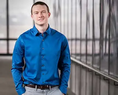 A person in a blue shirt and gray pants stands confidently in a modern building corridor with large windows and a blurred city view in the background, smiling and looking toward the camera.