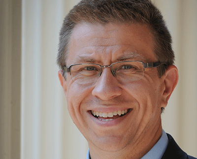 A person with short hair and glasses is smiling while looking at the camera. The background features vertical light-colored columns.