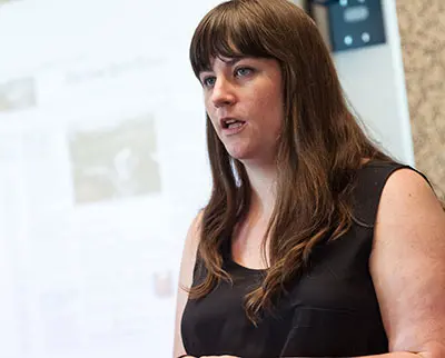 A woman with long brown hair and a black sleeveless top speaks in front of a blurred presentation screen. She appears focused and engaged in her topic.