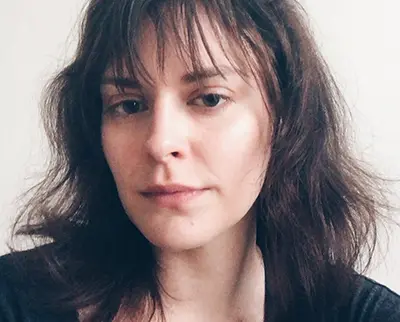 A person with long, brown hair and bangs is looking at the camera. They are wearing a dark top against a plain background.