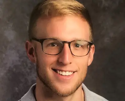 A man with short blond hair and glasses is smiling against a dark, textured background. He is wearing a light gray collared shirt.