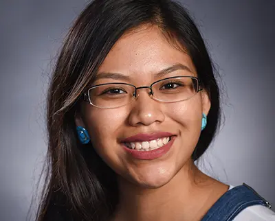 A young woman with long black hair and glasses smiles at the camera. She is wearing a blue top and blue flower-shaped earrings against a neutral gray background.