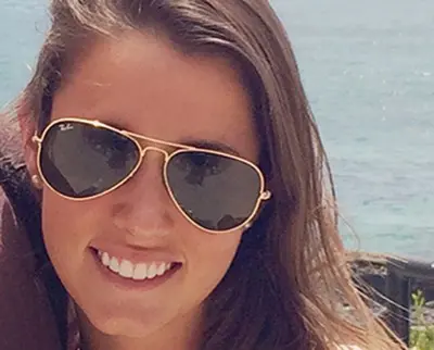 A person smiling while wearing aviator sunglasses, with long brown hair, against a backdrop of a body of water and clear skies.