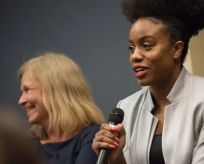 Two women are engaged in a discussion. The woman on the right, with an afro hairstyle, is speaking into a microphone and wearing a light gray jacket. The woman on the left, with blonde hair, is smiling and wearing a dark shirt.