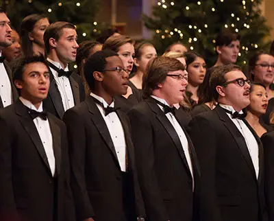 A choir of diverse individuals in black tuxedos sings in a festive setting, with decorated Christmas trees and lights in the background.