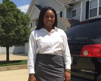 A person standing outside in front of a black car, wearing a white shirt and gray skirt. There's a house and a tree in the background under a clear blue sky.