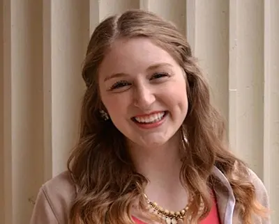 A young woman with long wavy hair is smiling warmly at the camera. She's wearing a necklace and a red top under a light-colored sweater. The background features vertical ridges in a neutral tone.