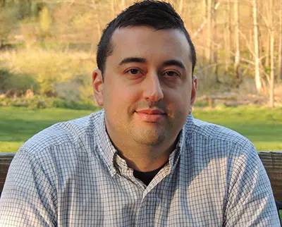 A person with short dark hair is sitting outdoors, wearing a checkered shirt. The background features a grassy area and trees with a soft light suggesting early morning or late afternoon.
