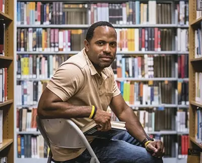 A person wearing a beige polo shirt and bracelets sits on a folding chair in a library. They are holding a book, surrounded by shelves filled with books. The atmosphere is calm and studious.