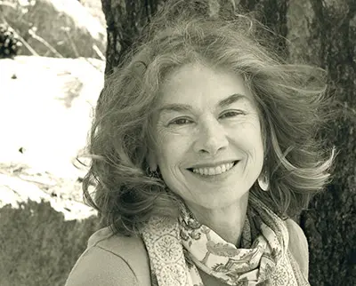 A person with wavy hair smiles while seated outdoors in front of a tree. They are wearing a patterned scarf and a light-colored top. The image is in sepia tone, adding a nostalgic feel to the scene.