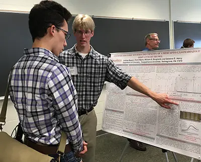 Two individuals in plaid shirts stand in front of a scientific poster presentation. One person gestures towards the poster, explaining to the other. A third person is blurred in the background, near a chalkboard.
