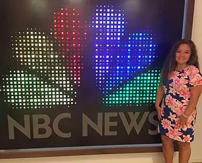 A person in a floral dress stands smiling next to the illuminated NBC News logo, which consists of colored dots forming a peacock shape. The background is dark, highlighting the vibrant colors of the logo.