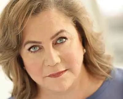 A woman with wavy, shoulder-length hair, wearing a blue top, looks directly at the camera. Her expression is calm and serious, with a soft focus background.