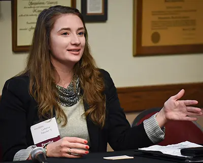 A woman with long brown hair speaks while gesturing with her hand. She sits at a table with a name tag and documents in front of her. Framed certificates hang on the wall behind.