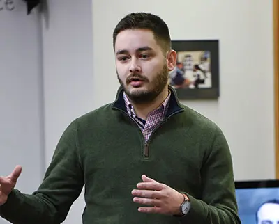 A man with short dark hair and a beard is speaking, gesturing with his hands. He is wearing a green pullover over a checkered shirt. A framed picture is visible on the wall behind him.
