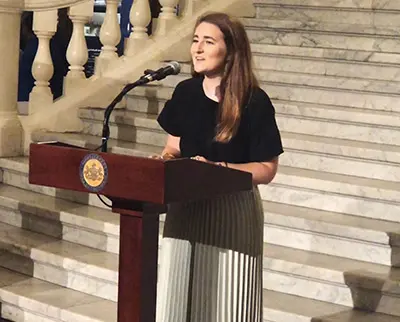 A person with long hair stands at a podium speaking into a microphone. They are on a marble staircase with ornate railings in the background, wearing a black top and a pleated skirt.