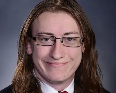 A person with long hair and glasses is smiling at the camera. They are wearing a suit and tie, and the background is a plain dark color.