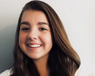 A young woman with long brown hair smiles at the camera against a neutral background. She is wearing a light-colored top.