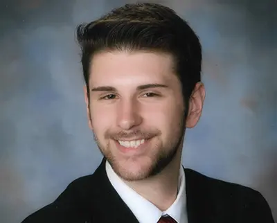 A person with short, dark hair and a beard is smiling in a formal portrait. They are wearing a black suit jacket, white shirt, and dark tie, set against a neutral blurred background.