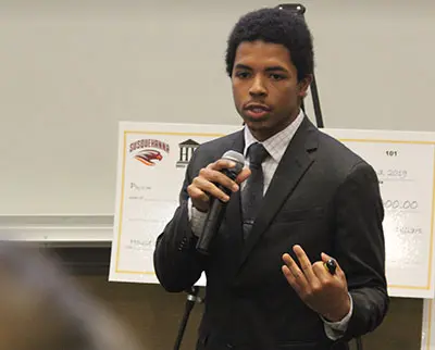 A person in a suit speaks into a microphone while holding a marker. They stand in front of a large check and presentation board.