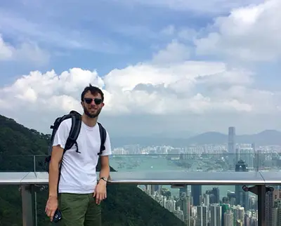 A person wearing sunglasses and a backpack stands on a viewing platform overlooking a cityscape with skyscrapers, mountains, and a bay under a partly cloudy sky.
