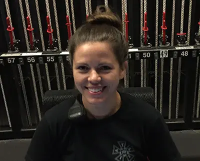 A person with a top-knot hairstyle is smiling in front of a backdrop of numbered ropes and red pulleys. They are wearing a black shirt and have a communication device on their shoulder.