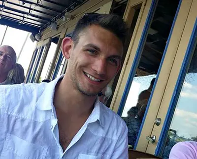 A man wearing a white shirt smiles at a camera while sitting outside on a patio. There are glass doors and a reflection of blue sky in the background, and another person is partially visible on the left.