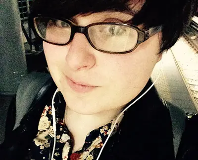 A person wearing glasses and earphones takes a selfie in a dimly lit setting. They have short dark hair and are wearing a floral-patterned shirt.