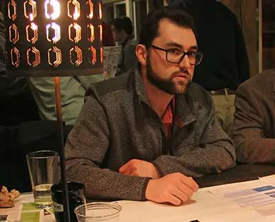 A man with glasses and a beard is seated at a table in a dimly lit setting. He is wearing a gray sweater and appears to be engaged in a conversation. On the table are papers, glasses, and a lamp emitting a warm light.