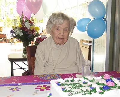 An elderly woman with curly white hair sits at a table decorated with a colorful cloth. There is a birthday cake with flowers and a message. Balloons and a bouquet of flowers are in the background.