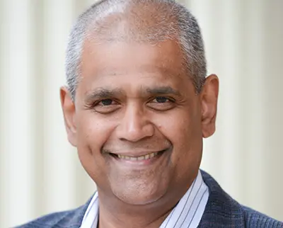 A person with short gray hair is smiling while wearing a striped shirt and a blazer. The background is a light, blurred setting.