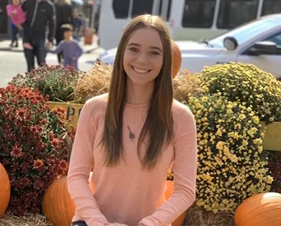 A smiling woman with long hair and a pink sweater sits among colorful autumn flowers and pumpkins. A street with a blurred background shows people and cars passing by.
