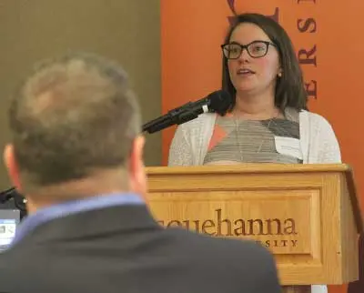 A woman in glasses speaks at a podium with a microphone. The podium has 