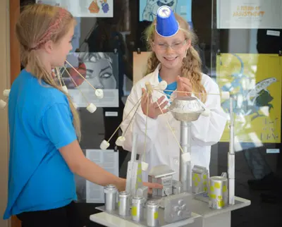 Two girls are smiling and interacting with a homemade science project made of cans, paper, and marshmallows. One wears a blue cone hat and glasses, and the other wears a blue shirt. There are posters on the wall behind them.