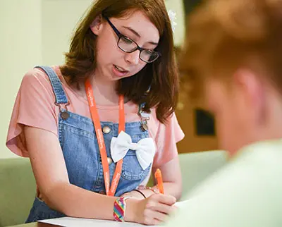 A young person with glasses, wearing a pink shirt and denim overalls, writes on paper at a desk. They have an orange lanyard and a white bow. Another person is blurred in the foreground.