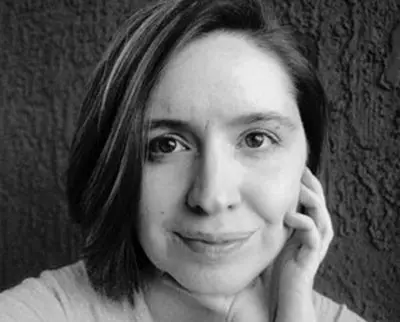 Black and white photo of a person with short hair smiling softly, resting their chin on their hand. The background is a textured wall.