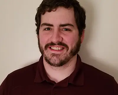 A person with short, dark hair and a beard smiles at the camera. They are wearing a maroon collared shirt. The background is plain and light-colored.