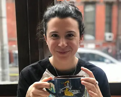 A person with dark hair is sitting indoors, holding a large cup with a colorful design. They are smiling softly. A window with a view of a building and part of a car can be seen in the background.