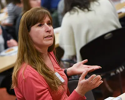 A woman with long brown hair, wearing a pink cardigan, gestures with her hands while speaking during a meeting. She is seated among other people at tables, appearing engaged in the discussion.