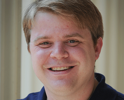 Smiling person with short, light brown hair wearing a dark shirt against a neutral background.
