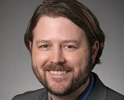 A man with brown hair and a beard, wearing a dark suit and light blue shirt, smiles against a gray background.